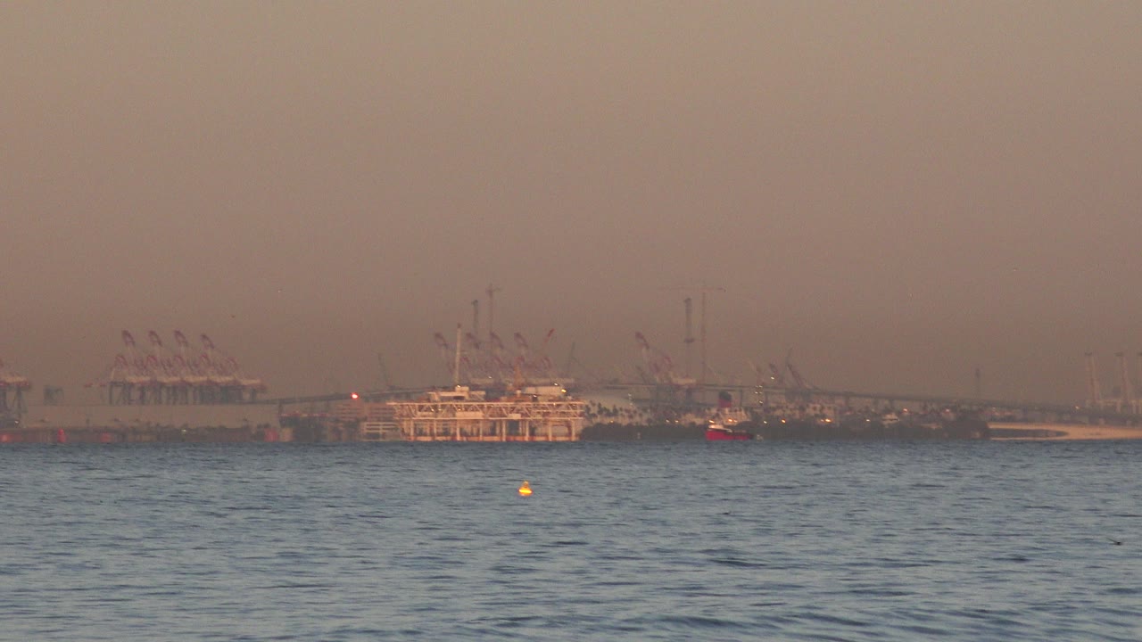 Huntington Beach ocean view at sunset with rippled blue water, a distant moored ship or platform, harbor cranes, and no visible people.