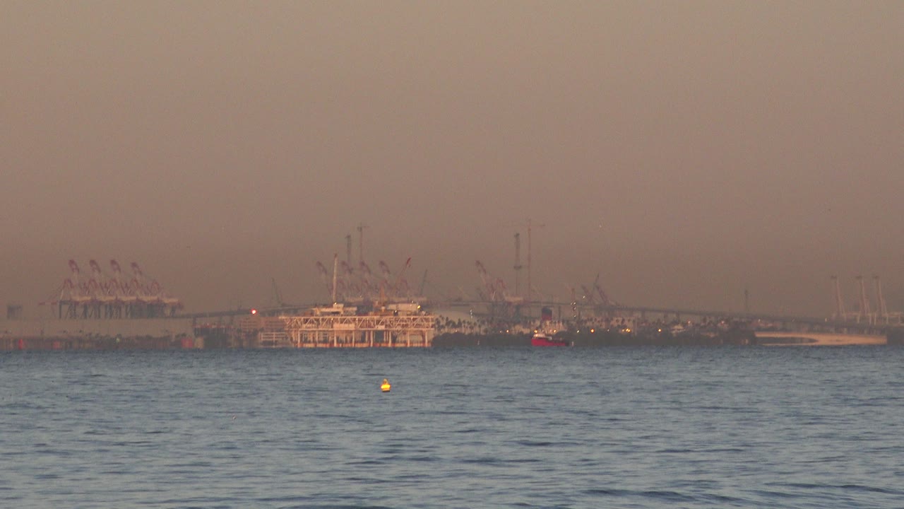 Huntington Beach water fills the lower frame while a distant moored vessel, cranes, and small boats sit under orange sunset haze with no visible people.