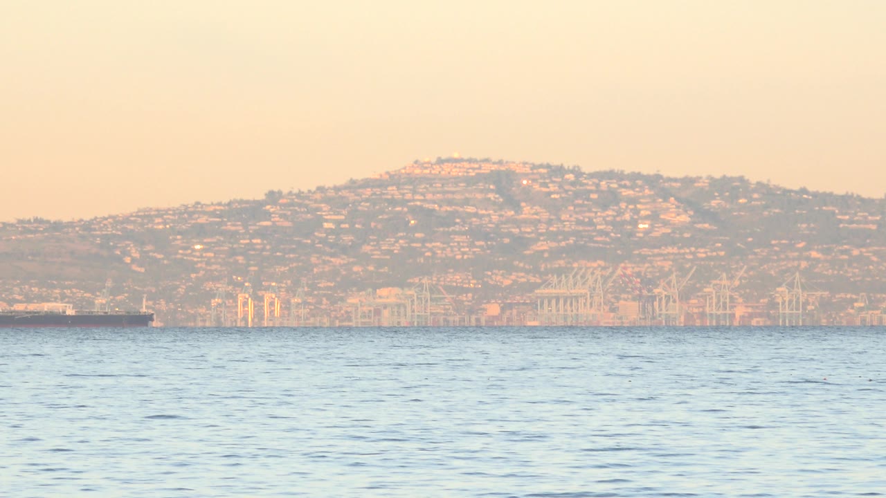 At 0:01, the ocean surface sits under warm hazy light with the Huntington Beach distant hillside and shoreline structures in the background, with no visible people.