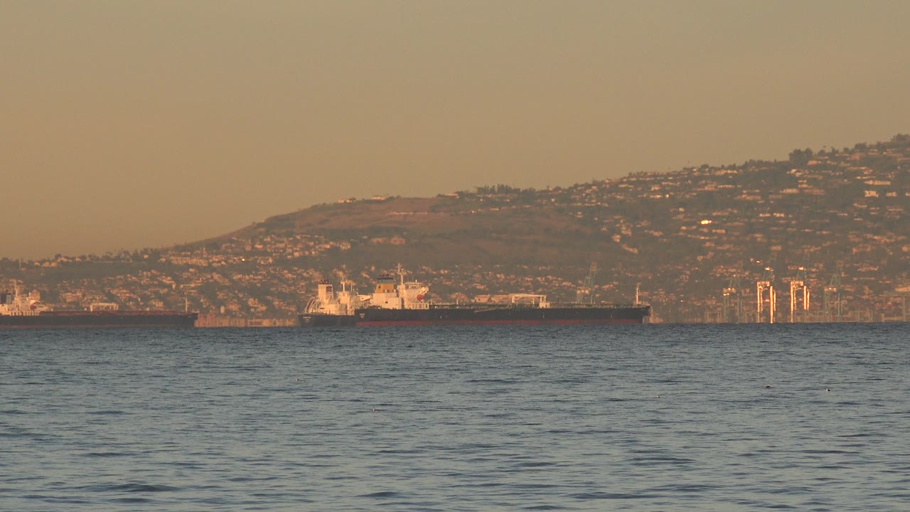 Ocean water with a distant cargo ship, harbor structures, and hillside buildings.