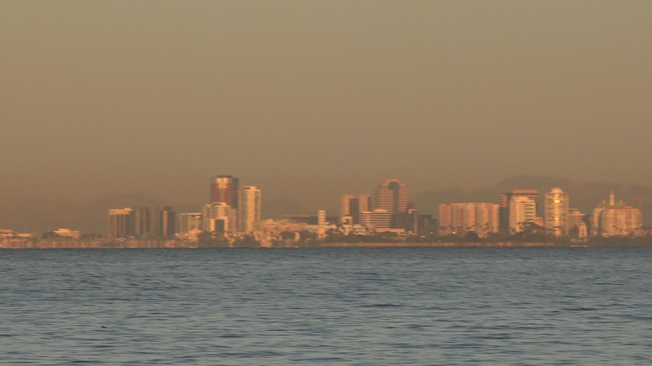 Opening frame of the Huntington Beach skyline seen far across blue rippled water, with warm haze over the buildings and no visible people.