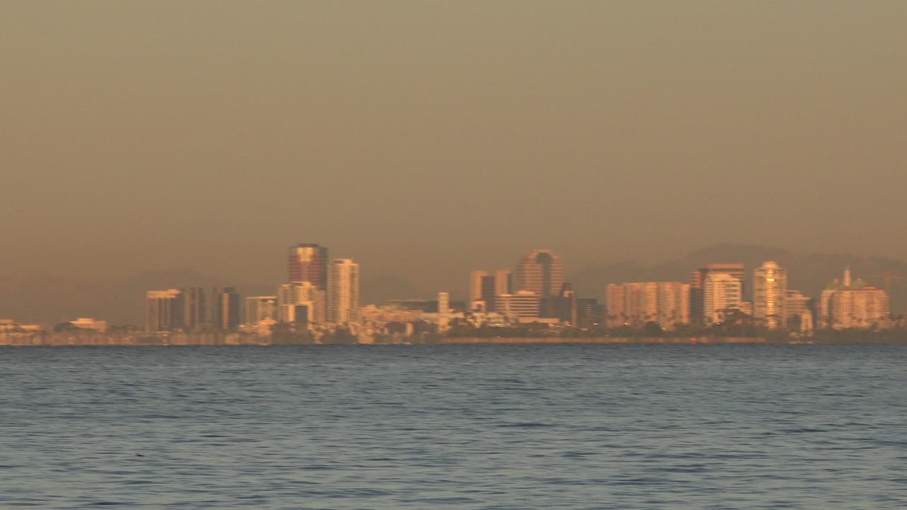 Huntington Beach buildings sit as a narrow distant band above calm rippled water, with hazy warm sky and no visible people.