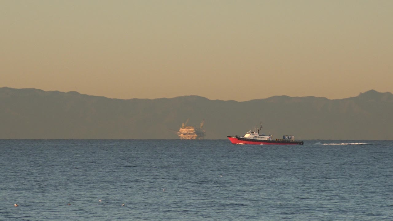 At 0:00 in Huntington Beach, California, a red-and-white boat sits left of center on blue water, with distant vessels near the horizon, hazy mountains behind them, and no visible people.