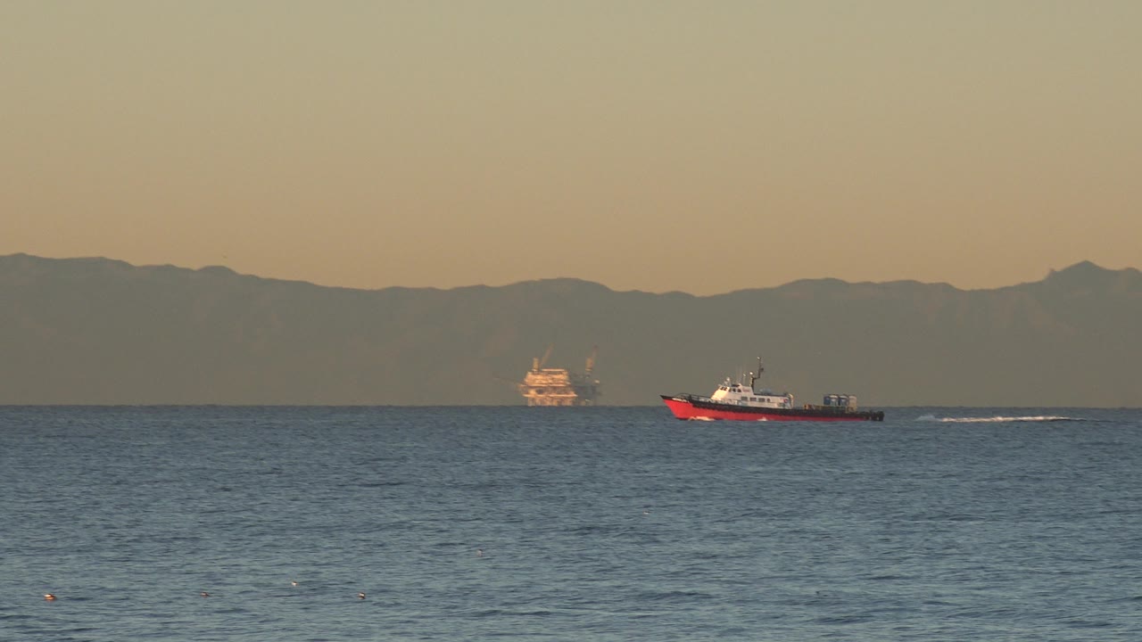 At 0:01 in Huntington Beach, California, the red-and-white boat moves across the midground with a thin wake trailing behind it, distant vessels on the horizon, hazy mountains, and no visible people.