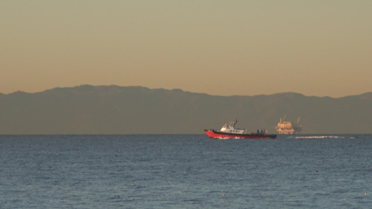 Red boat crossing open water off Huntington Beach, California, with a pale wake, distant hills, warm sky, and no visible people.