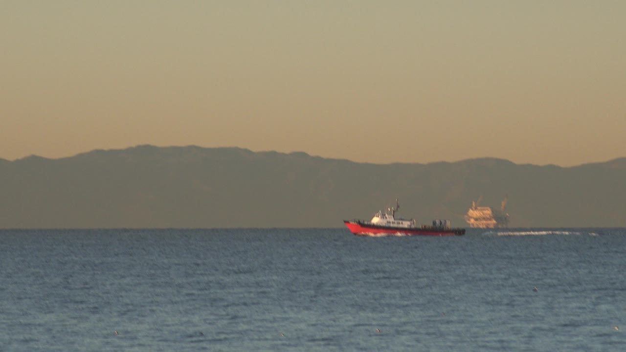 Red boat farther across the Huntington Beach ocean view, trailing a white wake beneath an amber sky with no visible people.