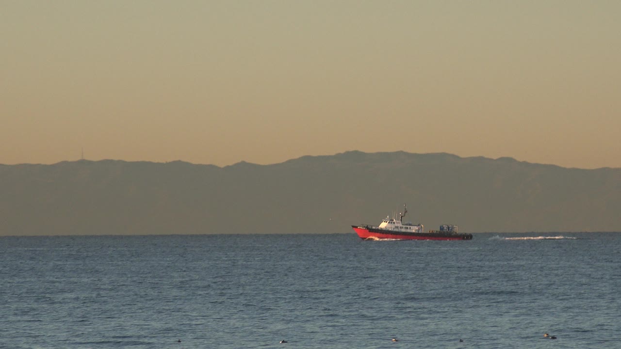 At 0:00 in Huntington Beach, a small red and white boat sits left of center on blue ocean water with a hazy mountain ridge behind it and no visible people.
