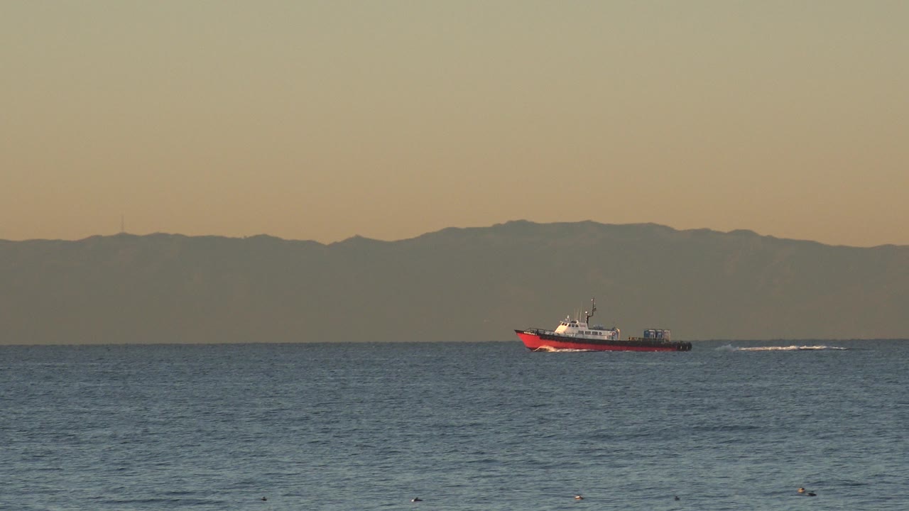 At 0:01 in Huntington Beach, the red and white boat appears farther across the frame with a thin white wake trailing over the blue water and no visible people.
