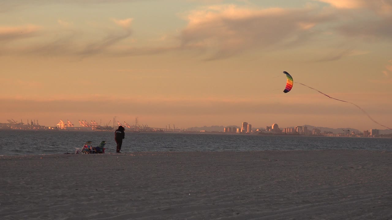 Wide Huntington Beach frame showing open sand, a distant person near the waterline, beach chairs or gear, a colorful kite overhead, and the city skyline across the horizon.