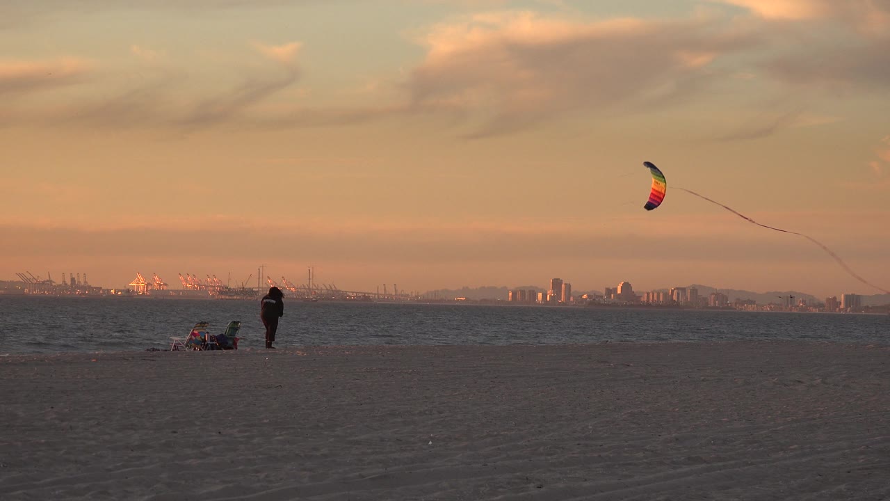 Huntington Beach view with the kite higher in the sky, a person standing on the sand, seated beach gear nearby, low waves, and port-like structures on the far horizon.