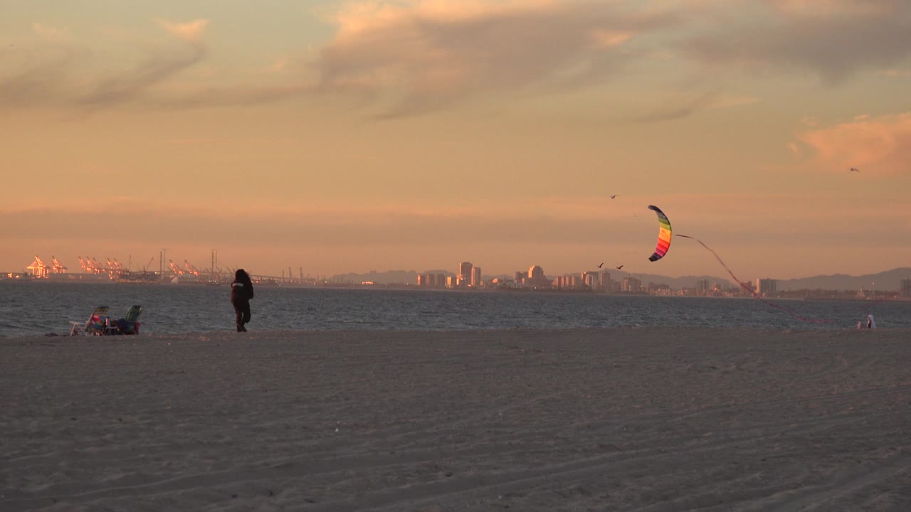 Wide Huntington Beach frame with a dark-clothed person flying a multicolored kite over open sand, with ocean water, distant shoreline structures, birds, and warm sky behind them.