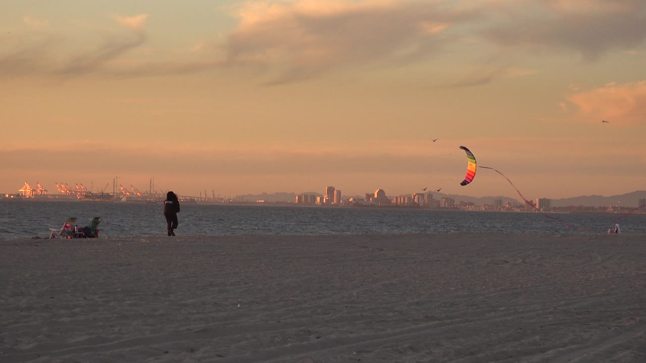 Huntington Beach view one second in, with the kite arcing above the ocean while the person remains on the sand near seated beachgoers and the distant shoreline sits under orange light.
