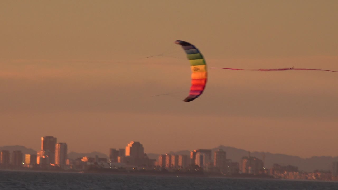 The rainbow kite sits near the center of the Huntington Beach sky, with its tail stretched sideways over dark water and distant buildings.