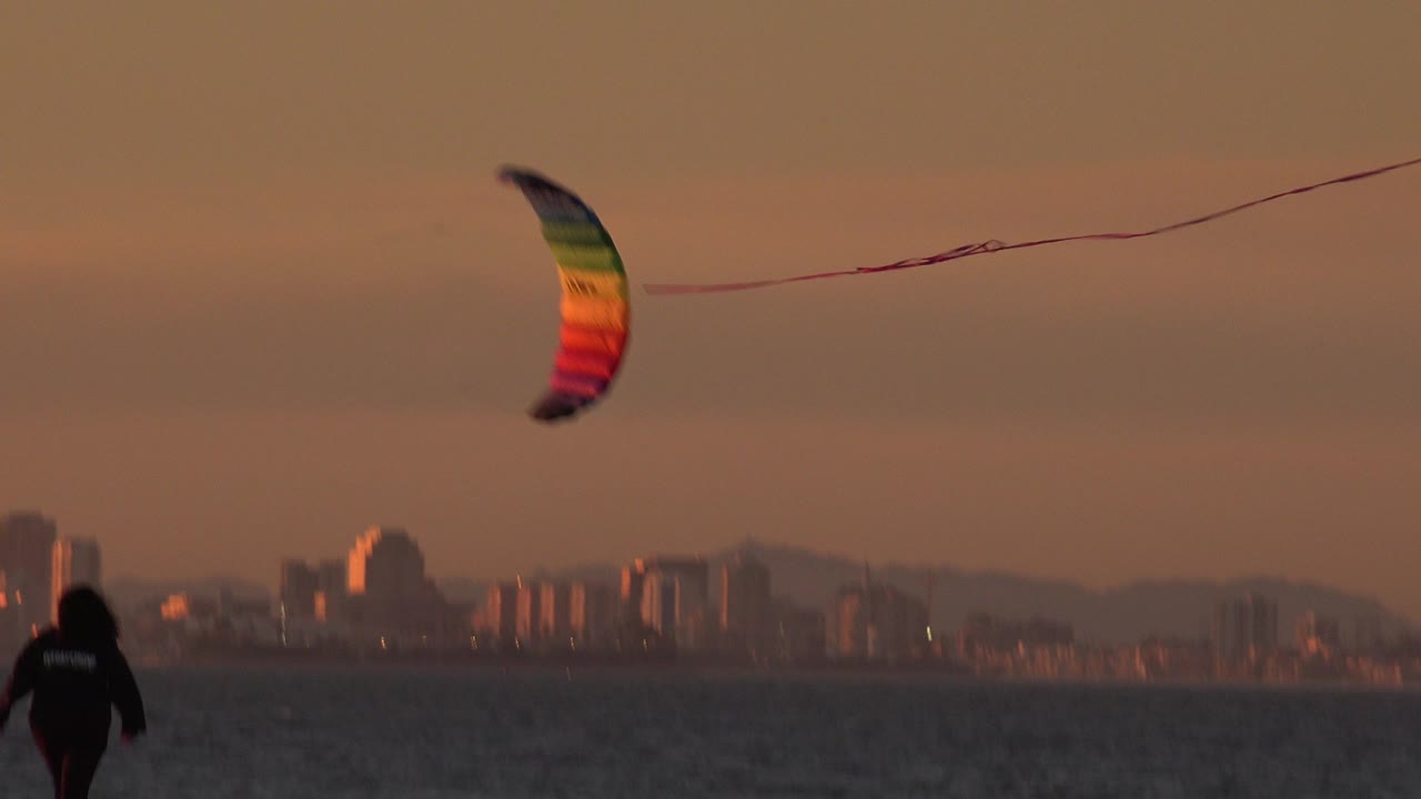 Rainbow kite over ocean with a beach walker and distant skyline at twilight.