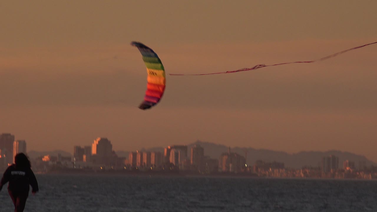 The rainbow kite stays above the ocean while the beach walker appears near the lower center of the Huntington Beach frame, backed by a hazy city skyline.