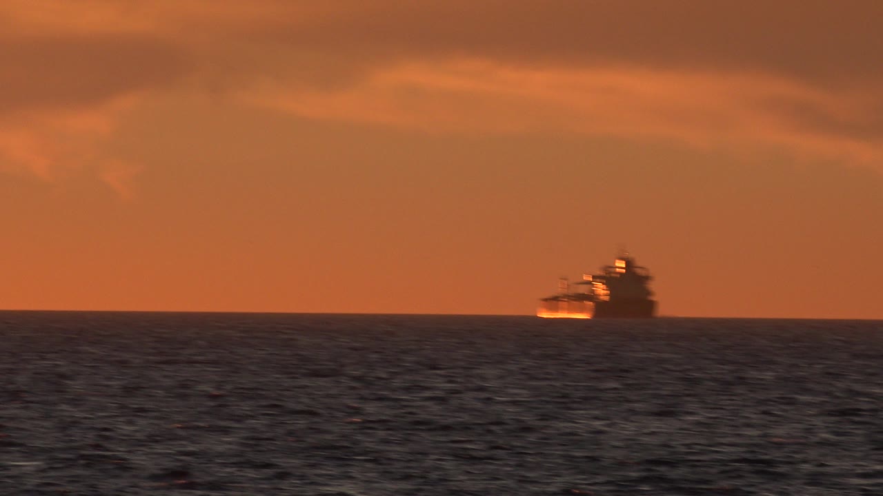 A distant large vessel sits near the horizon off Huntington Beach, California, with dark water in the foreground, orange clouded sky above, and no visible people.