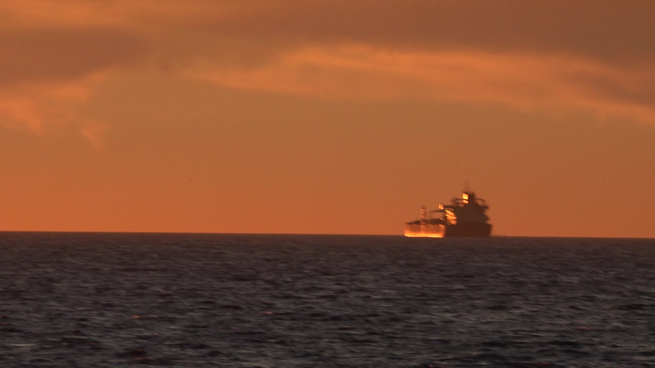 The vessel remains near the center of the horizon at Huntington Beach, California, with choppy dark water below a warm orange sky and no visible people.