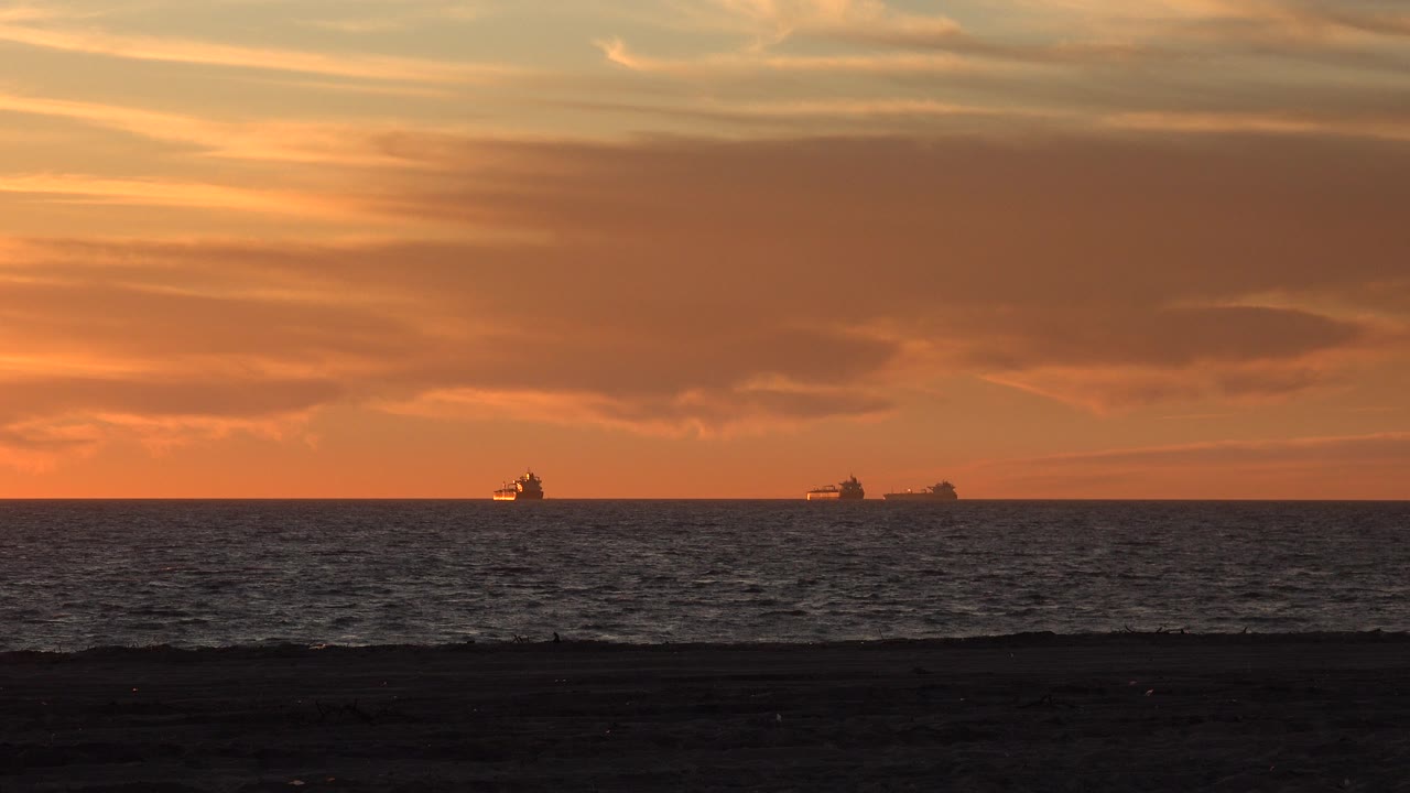 Huntington Beach sunset view with a dark beach foreground, rippled ocean, distant ship silhouettes near the horizon, orange clouds, and no visible people.