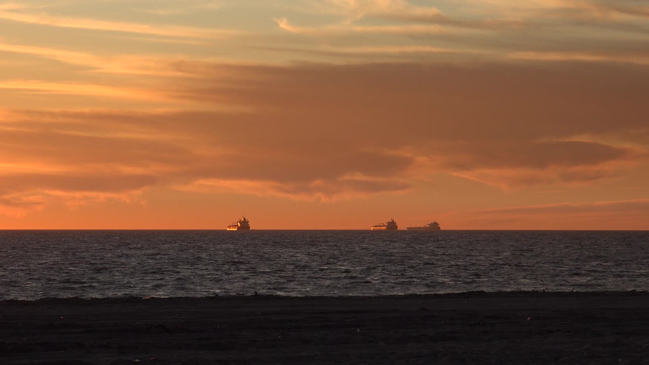 Huntington Beach ocean horizon at sunset with small ship silhouettes separated across the water, a dark strip of beach, orange cloud layers, and no visible people.