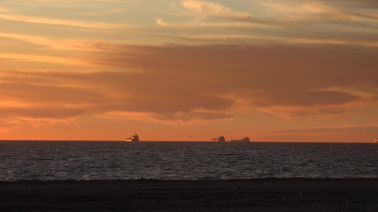 Huntington Beach sunset frame with orange clouds, a dark ocean band, two small ships near the horizon, and tiny shoreline silhouettes in the foreground.