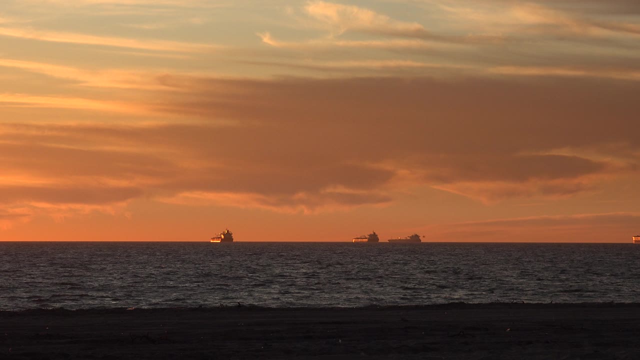 Orange sunset and dark ocean with small ships beyond the beach.