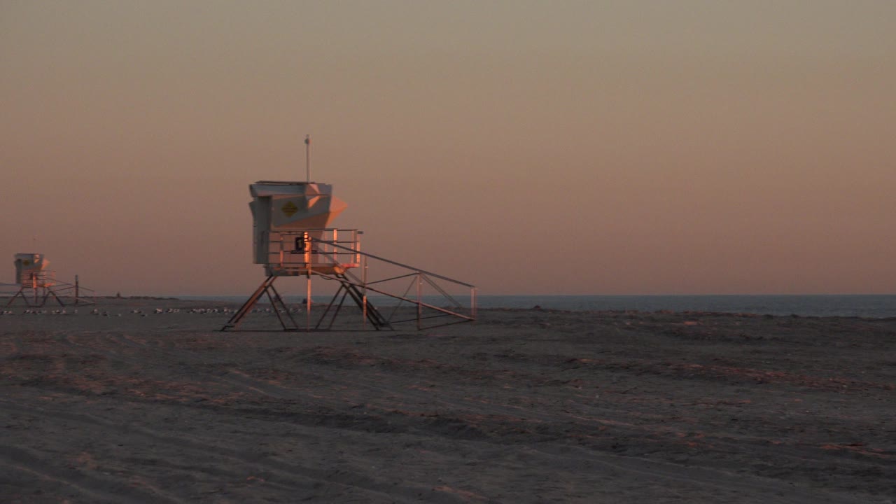 Wide Huntington Beach sunset frame with a main lifeguard tower left of center, a smaller tower in the distance, ocean horizon behind them, and no visible people.