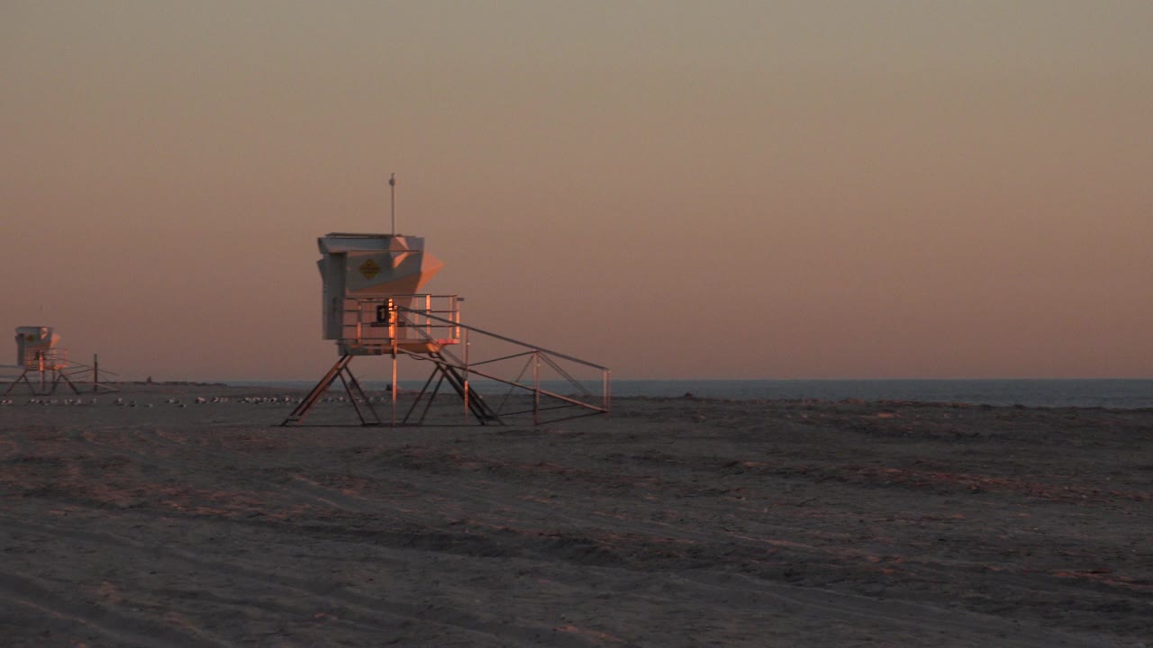 Huntington Beach dusk view with the main lifeguard tower near the center, a second tower farther back, dark sand foreground, ocean line, and no visible people.