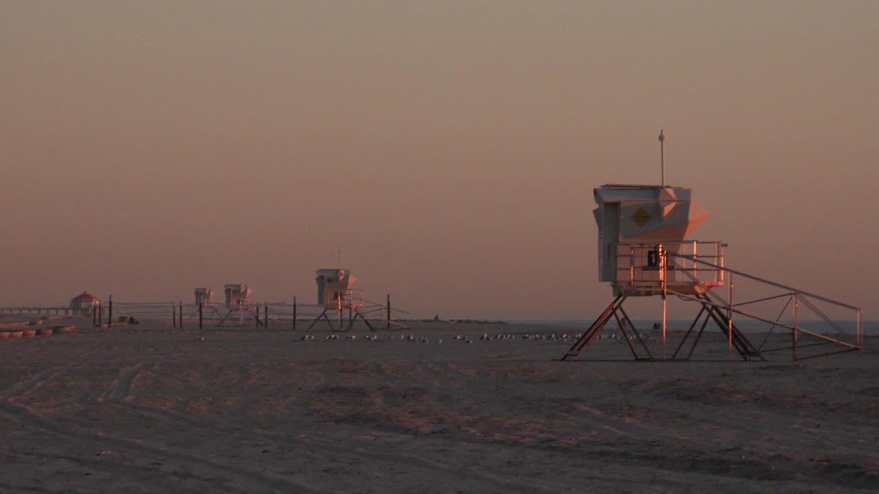Huntington Beach frame showing several elevated angular station structures across open sand under warm evening light, with no visible people.