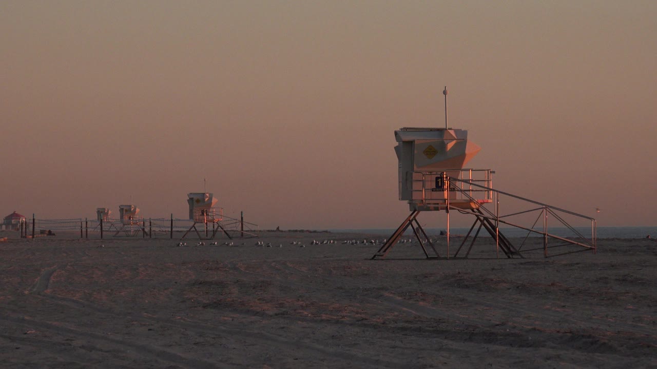 Huntington Beach sunset view with several lifeguard towers spaced across dark sand, a low ocean horizon, pink-red sky, and no visible people.