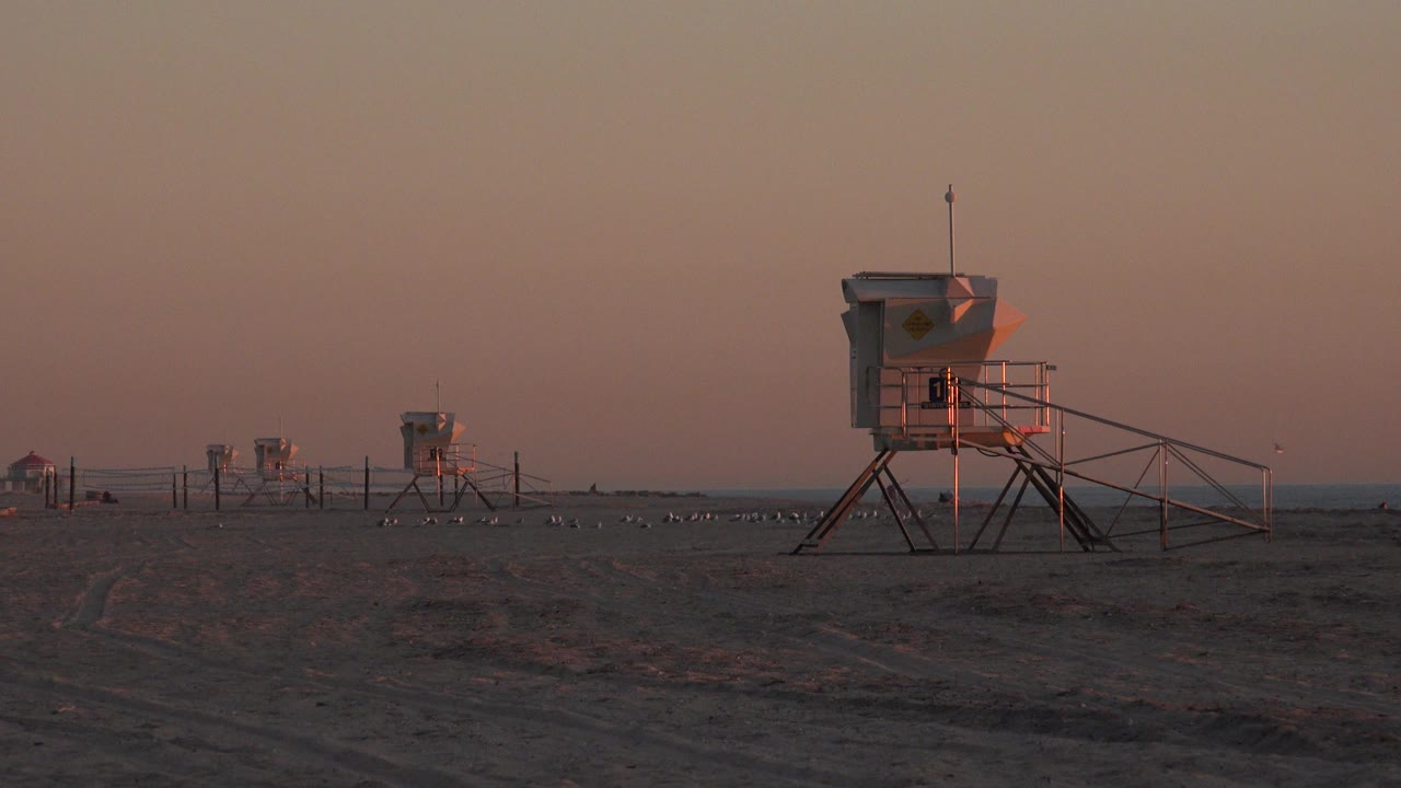 A foreground lifeguard tower stands right of center on Huntington Beach sand, with distant towers, a thin ocean line, warm sky color, and no visible people.