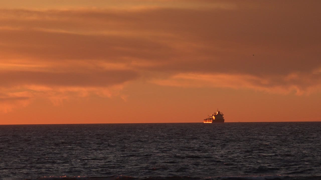 A distant passenger ship sits left of center on the Huntington Beach ocean horizon under orange clouds, with dark water in the foreground and no visible people.