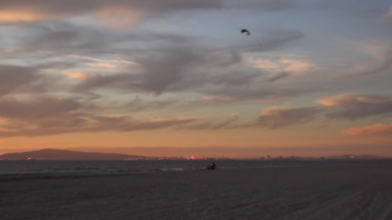 A small kite sits high over Huntington Beach at dusk, with dark sand in the foreground, low surf on the horizon, pastel clouds, and no visible people.