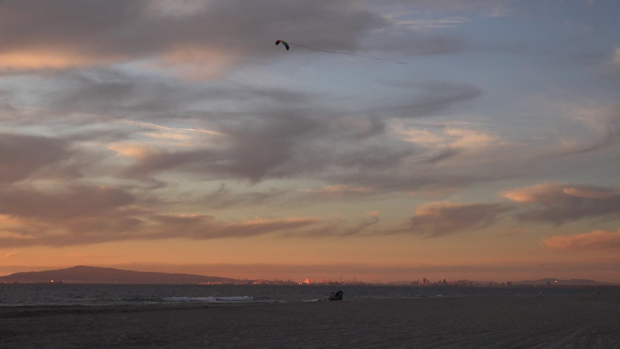 A small stunt kite flies high above Huntington Beach at sunset, with dark sand, low surf, distant hills, and orange-gray clouds filling the frame.