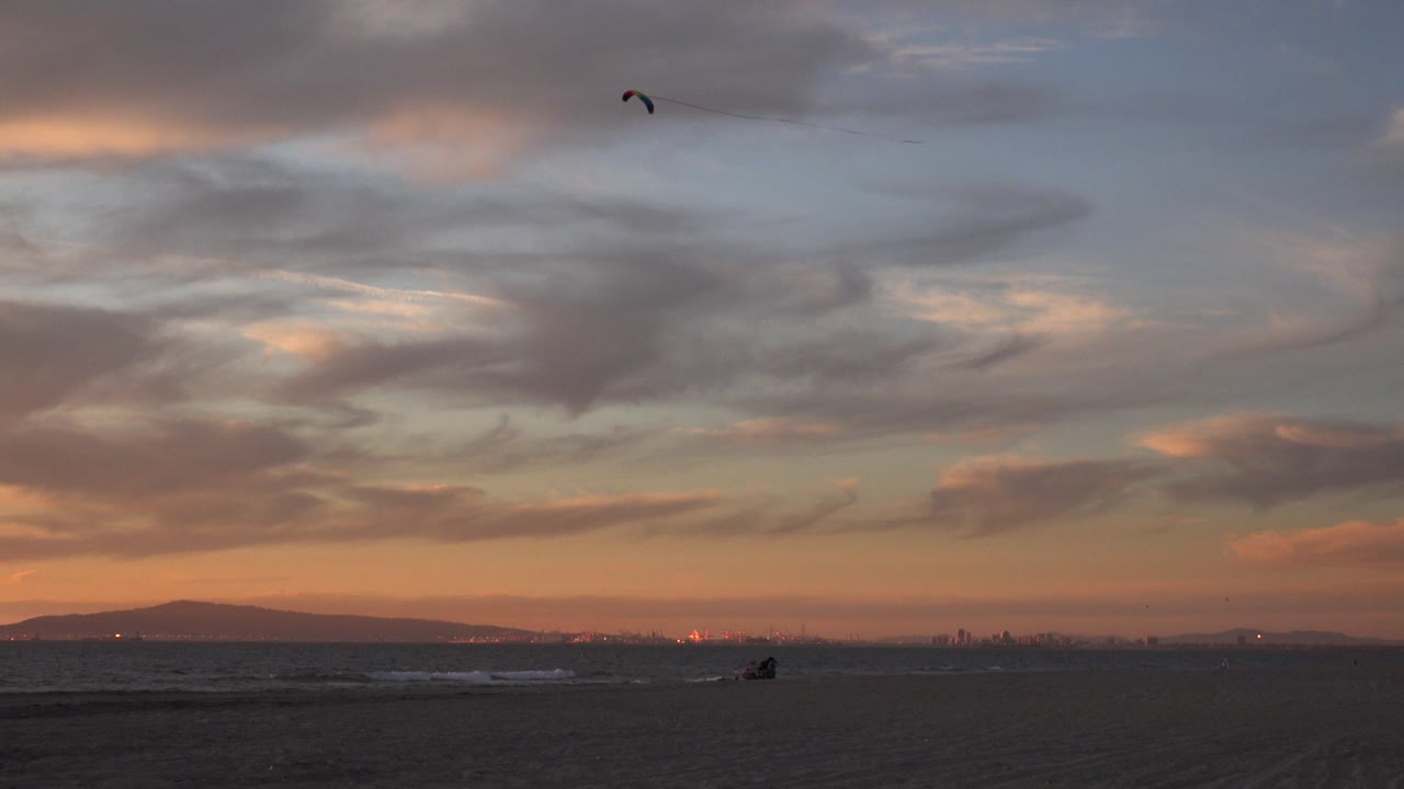 The stunt kite appears near the top of the Huntington Beach sky while the ocean horizon, dark beach, and warm sunset band remain low in the frame.