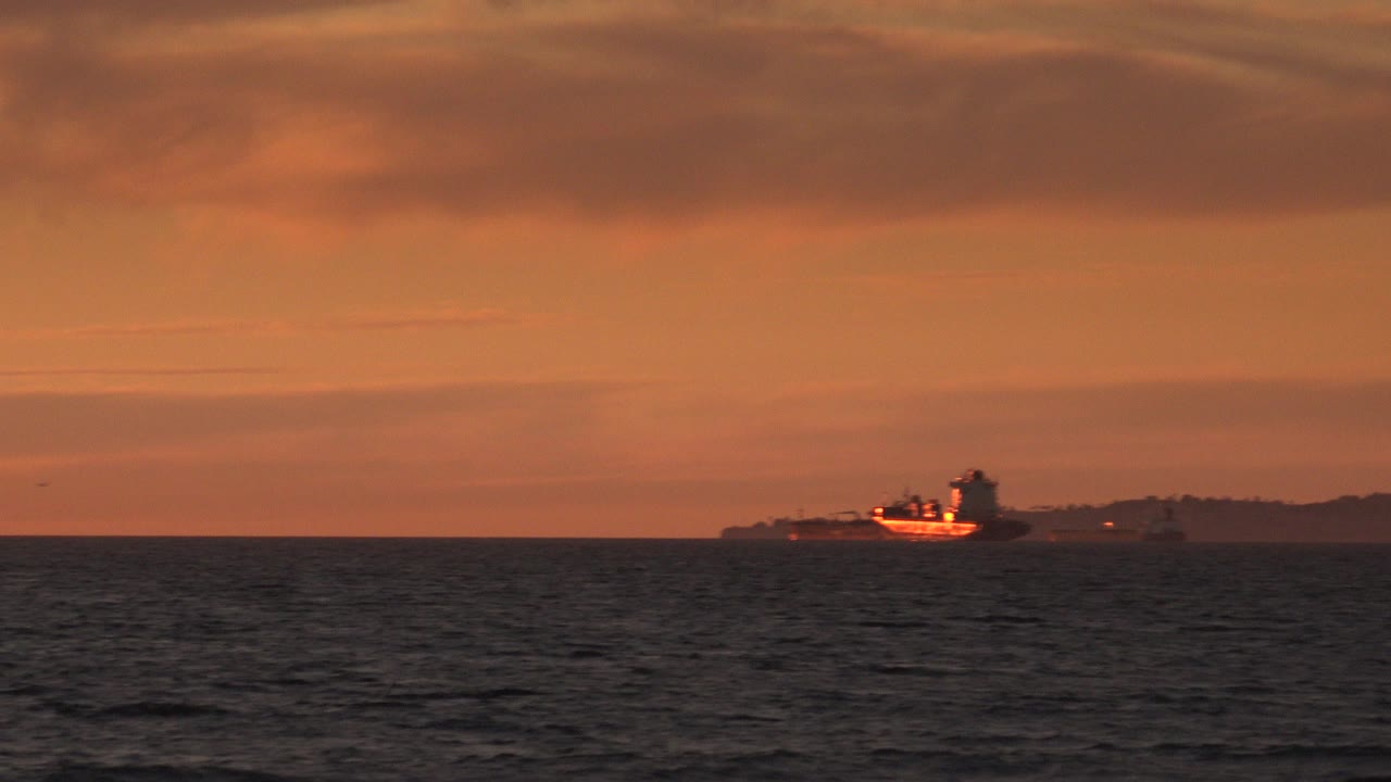 Distant tugboat on orange sunset water near a dark shoreline.