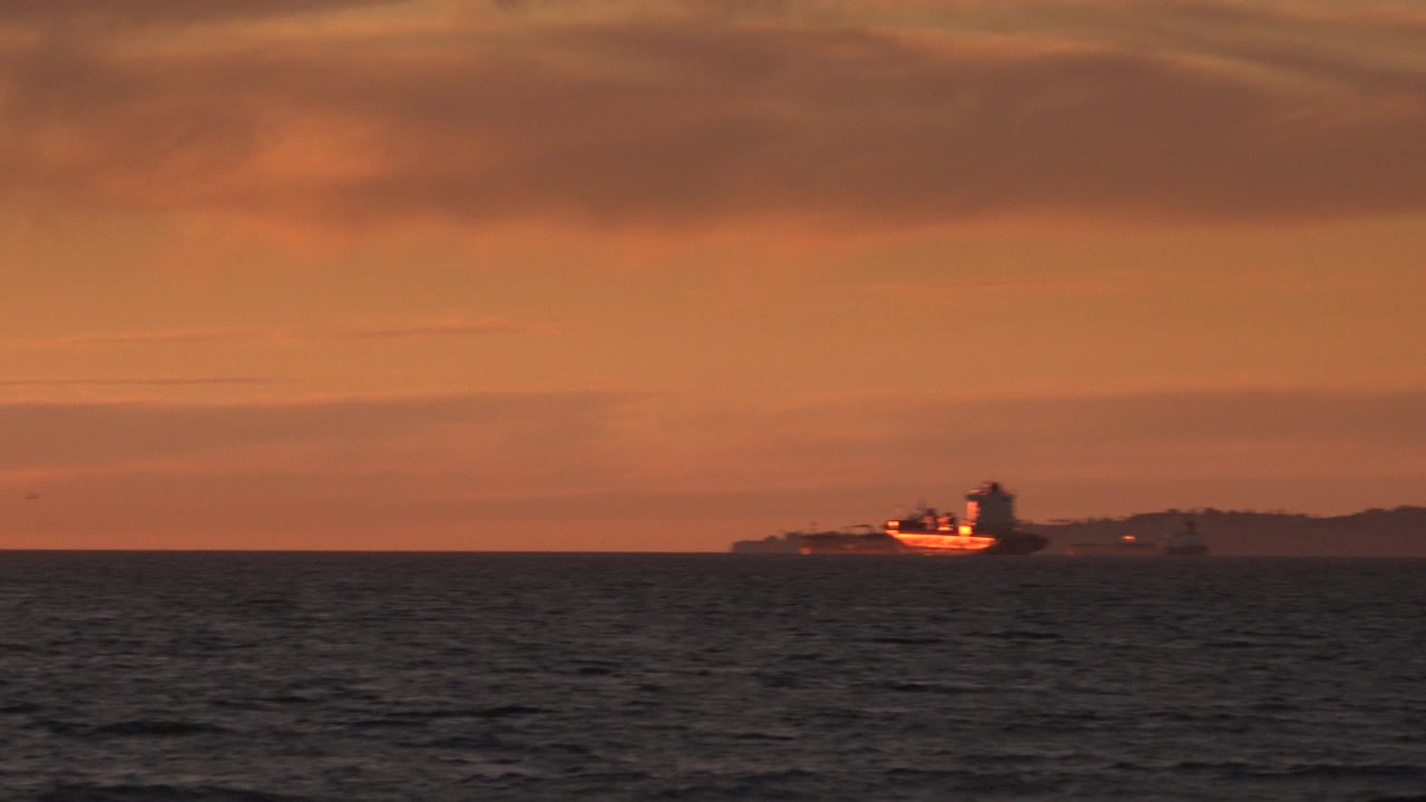 At 0:01, the tugboat remains distant near the horizon, its hull catching orange light while dark water fills the foreground, with no visible people.