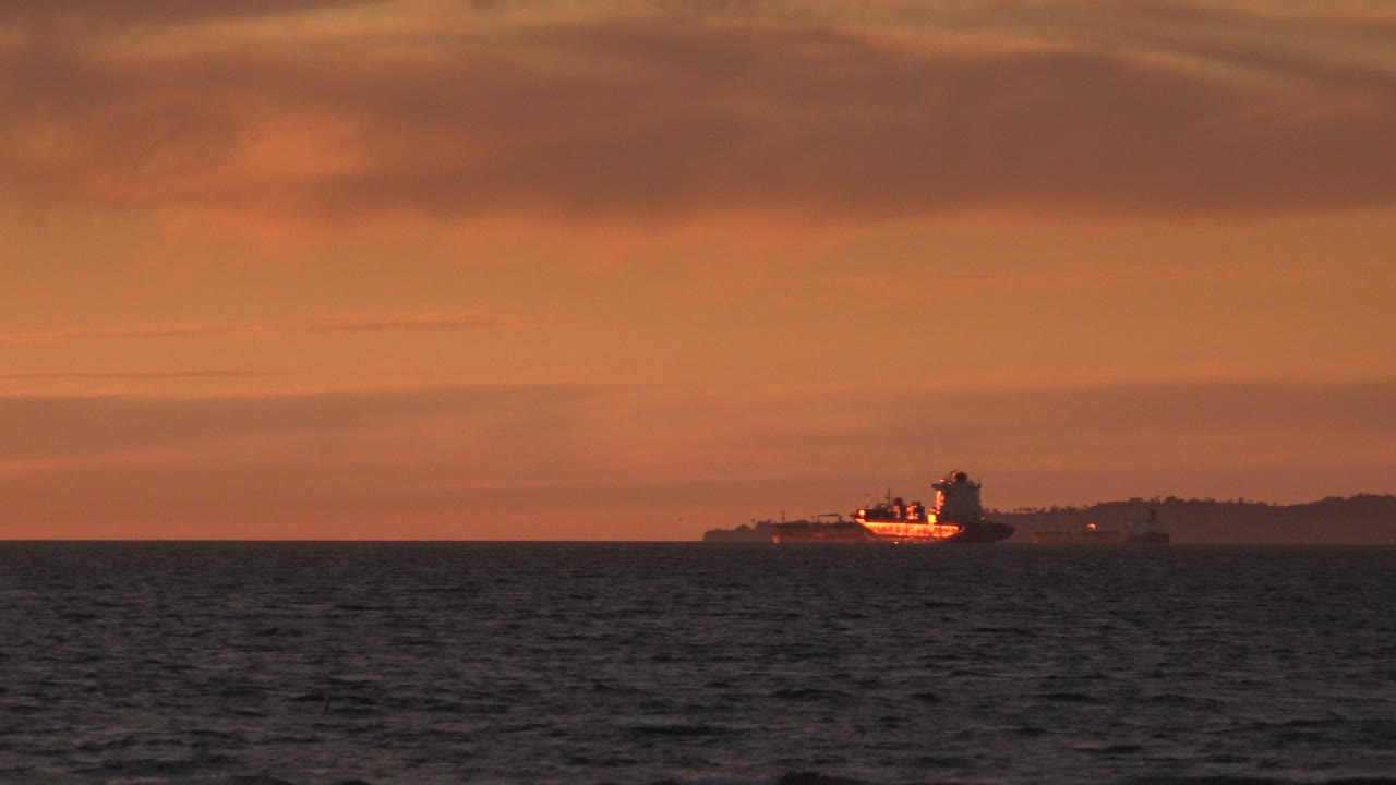 At Huntington Beach, California, a distant vessel sits near the horizon beyond dark ocean water under an orange sunset sky, with no visible people.