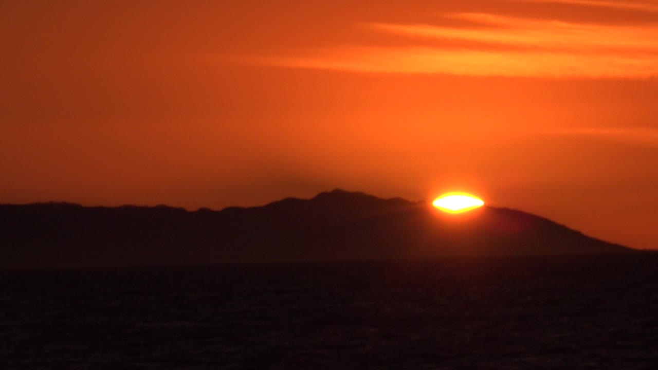 At 0:00 in Huntington Beach, California, the orange sun sits low behind a dark ridge across the ocean, with faint cloud bands above and no visible people.