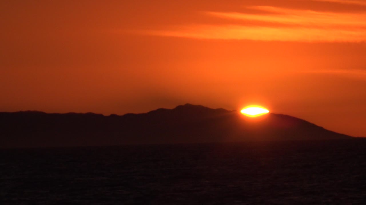 At 0:01 in Huntington Beach, California, the sun remains partly blocked by the silhouetted landform, with dark ocean texture below and no visible people.