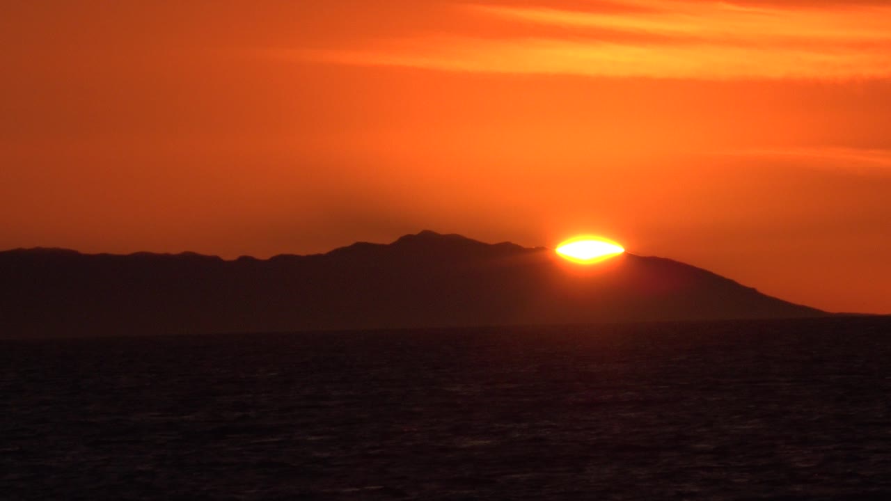 Bright sunset disk above a dark island silhouette and ocean.