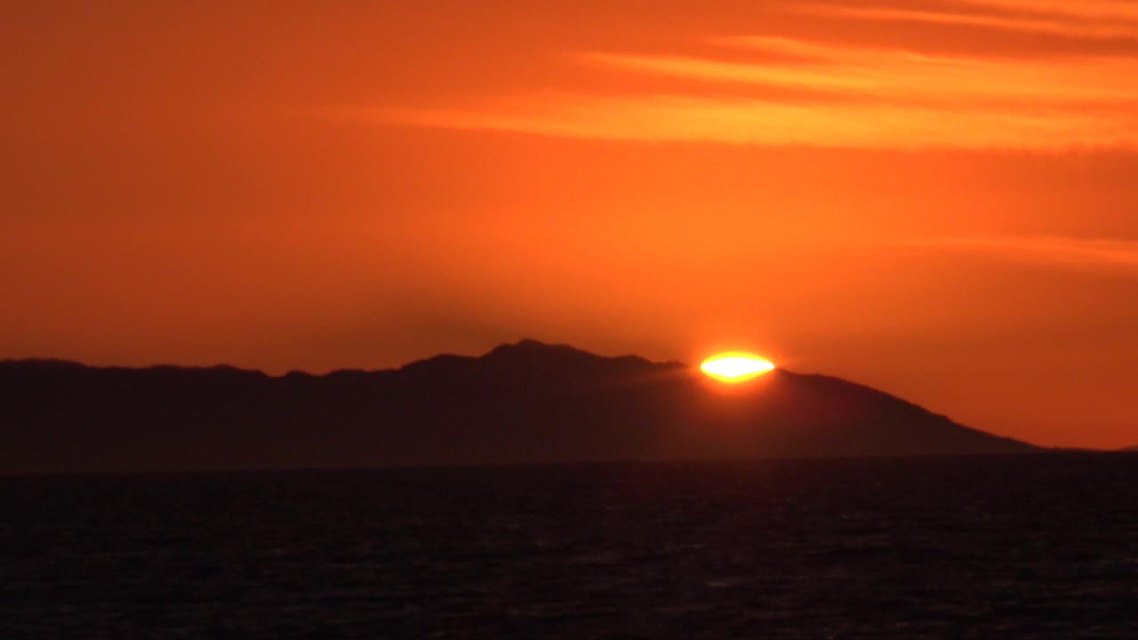 At 0:00 in Huntington Beach, California, an orange sun sits just above a dark mountain ridge with ocean in the foreground and no visible people.
