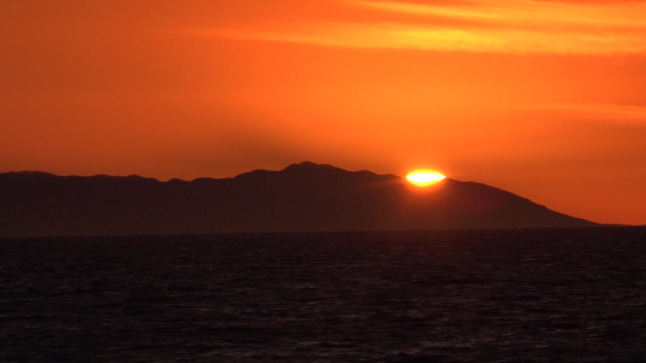 At 0:03 in Huntington Beach, California, the sun is lower against the ridge and the sky is saturated orange above a nearly black ocean foreground, with no visible people.