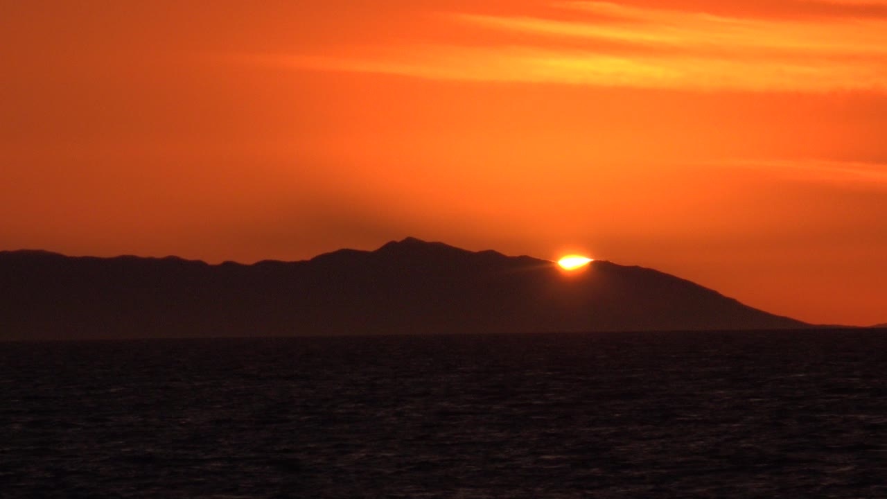Orange sunset over ocean with the sun touching a dark hill silhouette.