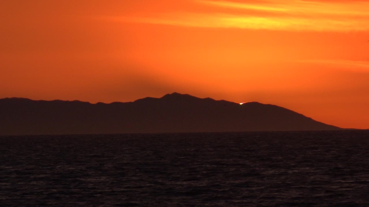 Huntington Beach sunset view with dark ocean in the foreground, silhouetted hills on the horizon, orange cloud bands above, and no visible people.