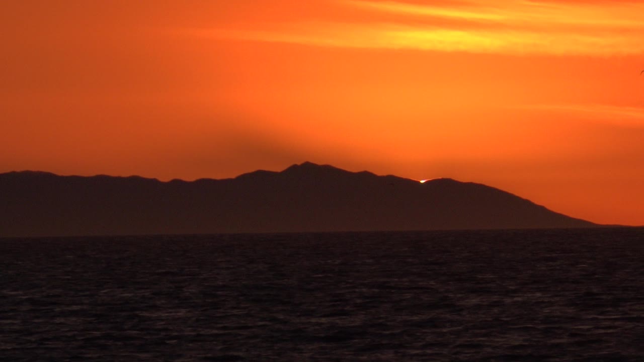 Huntington Beach coastal frame showing choppy dark water, a low silhouetted ridge, a small bright sunset glow near the hills, and no visible people.