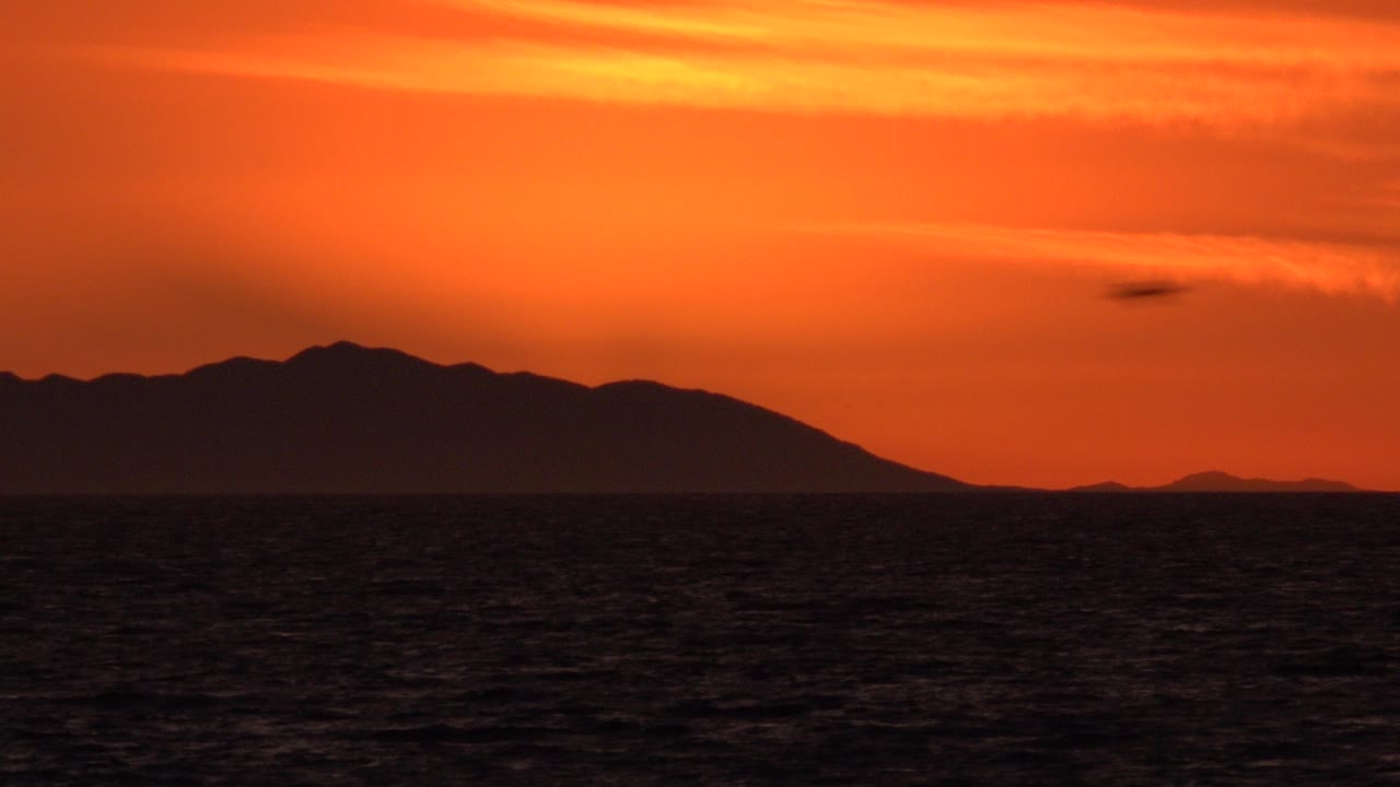 Huntington Beach, California sunset frame with dark ocean in front, a broad silhouetted landform across the horizon, orange cloud bands above, and no visible people.