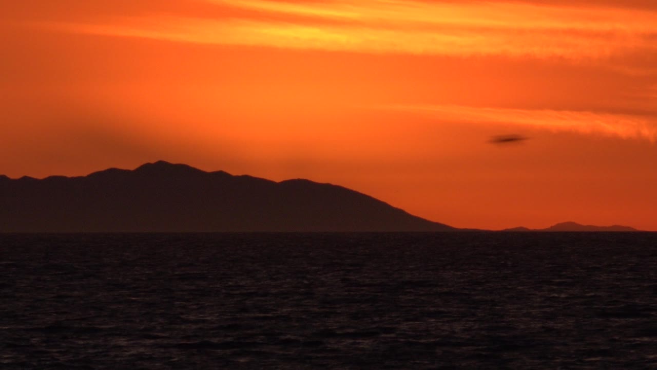 Huntington Beach, California sunset view with the silhouetted landform shifted across the center, dark water below, orange sky above, and no visible people.