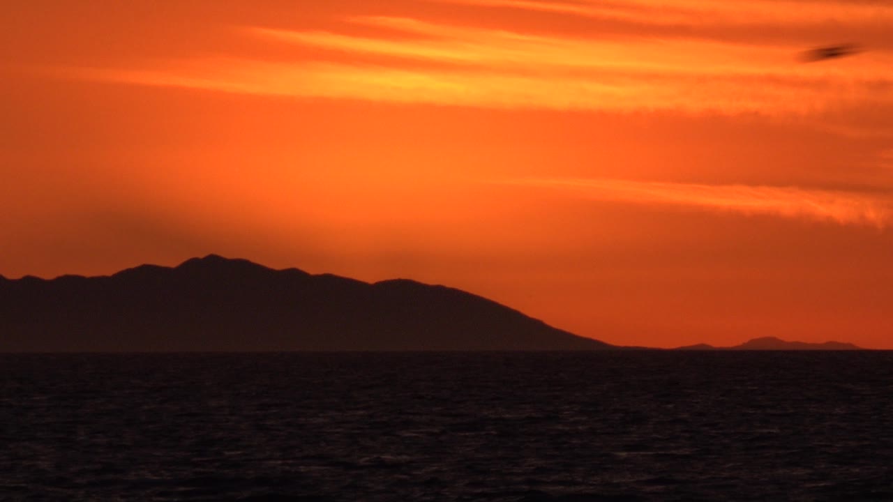 Dark island or mountain shape beyond ocean beneath orange evening clouds.