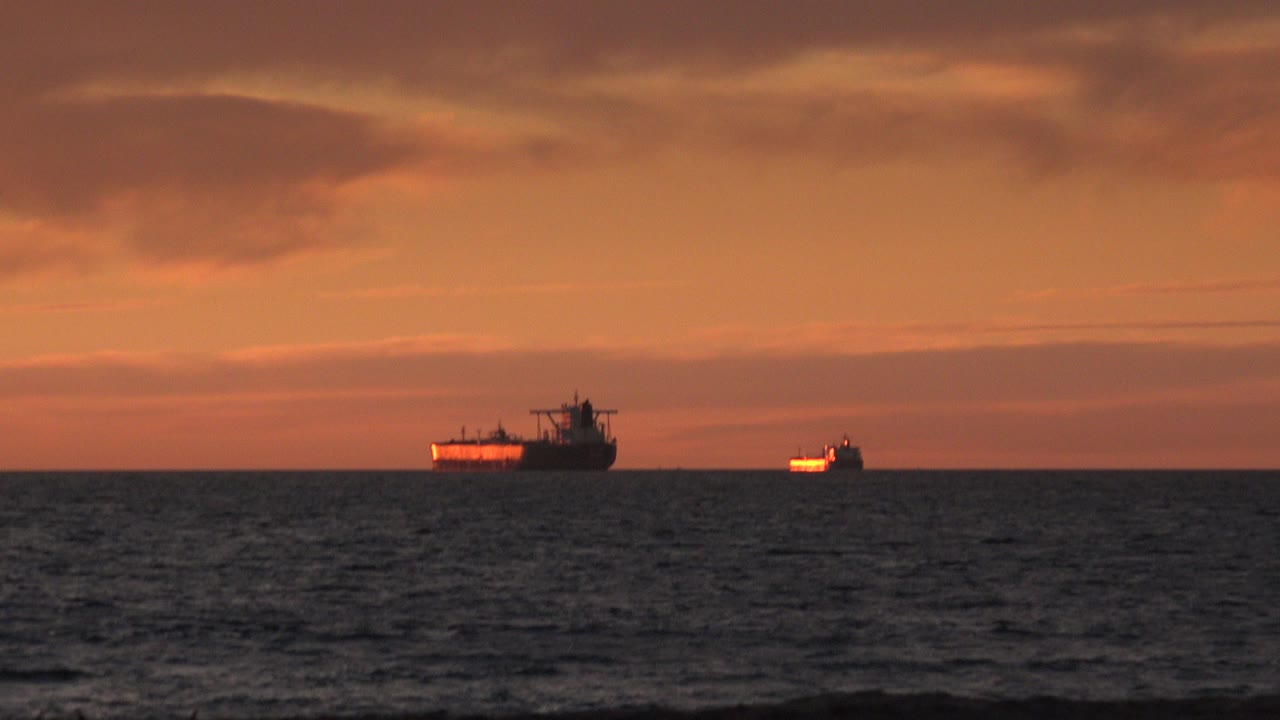 At Huntington Beach, two distant ships sit on the ocean horizon beneath orange evening clouds, with dark textured water in the foreground and no visible people.
