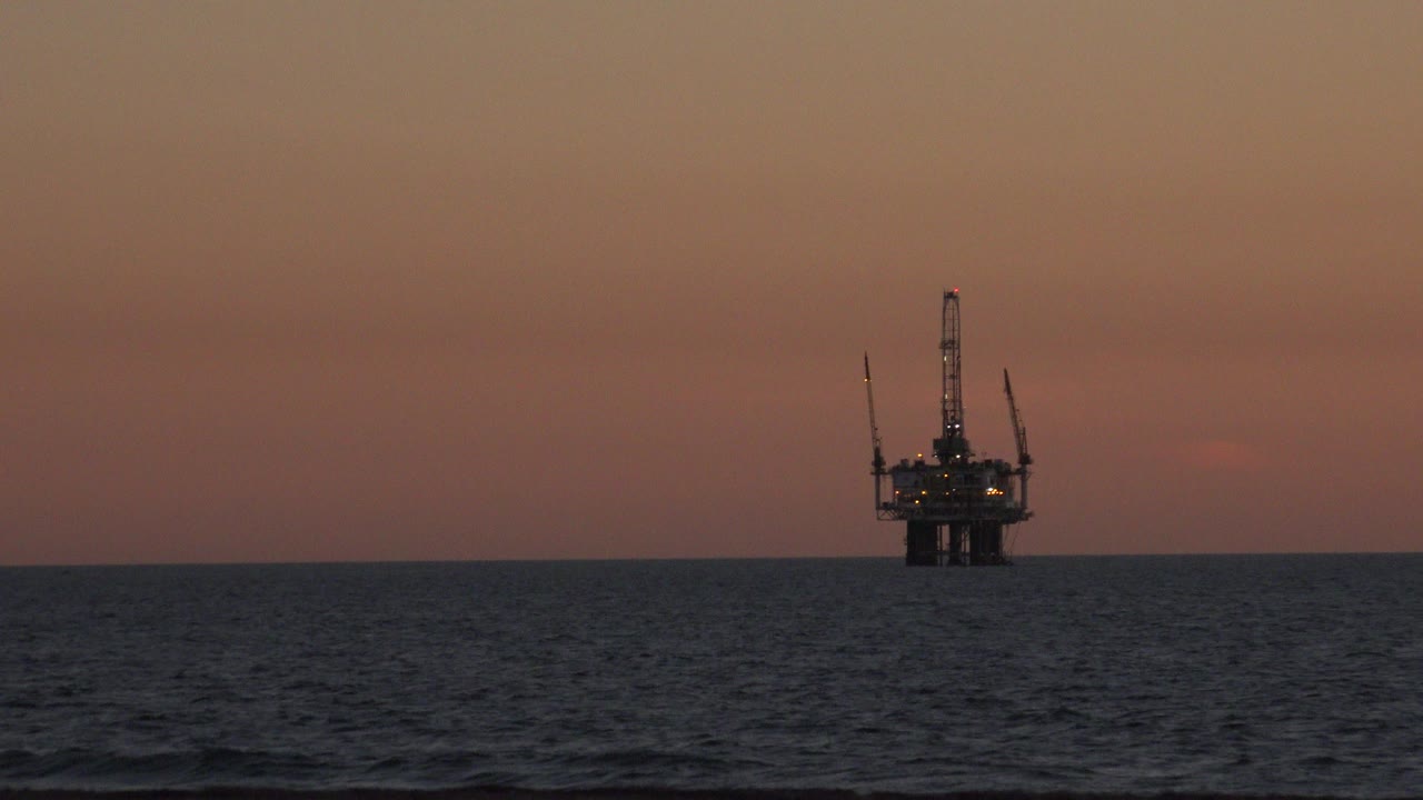 Distant offshore oil platform on the Huntington Beach horizon under an orange sunset sky, with dark ocean waves in front and no visible people.